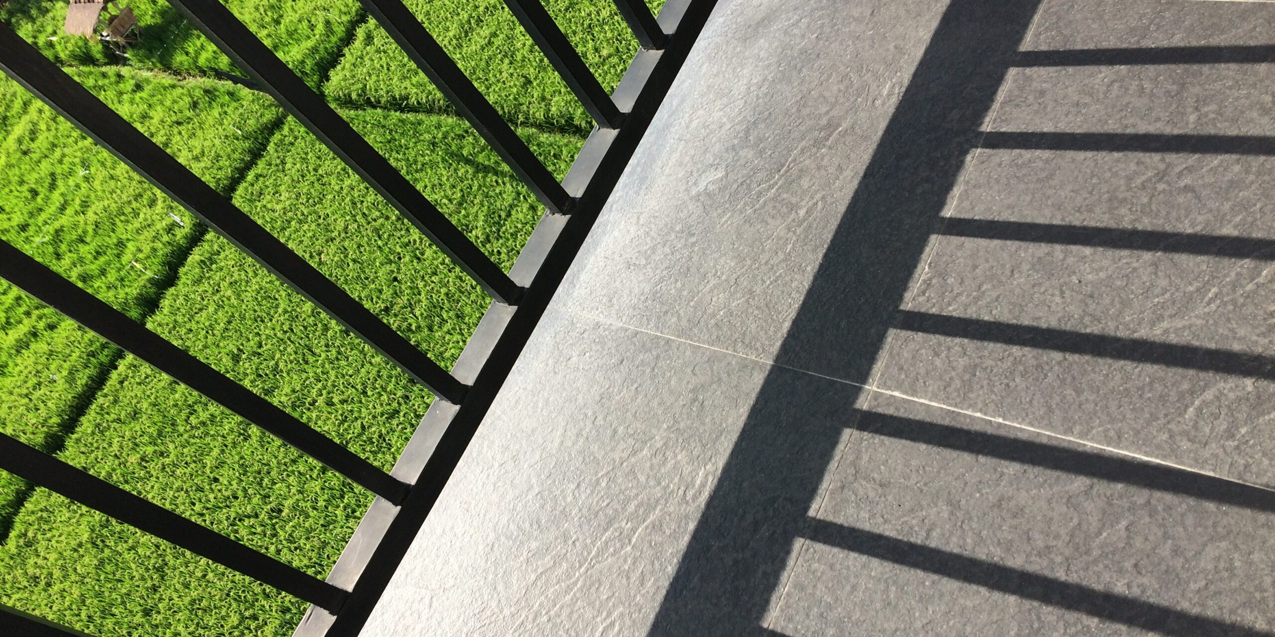 A metal fence shadow on the balcony with a view of a grassy field on a sunny day